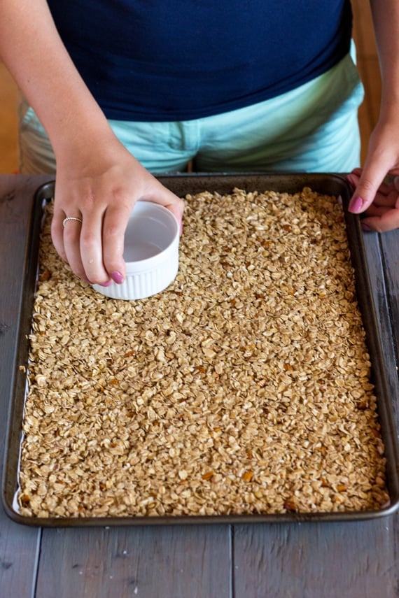 A woman is using a ramekin to distribute Crunchy Homemade Granola evenly throughout the pan