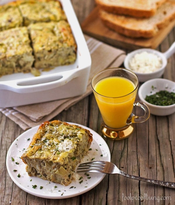  A serving of Eggs with Artichoke, Tarragon, and Parmesan with a glass of orange juice with a platter of the egg dish in the background