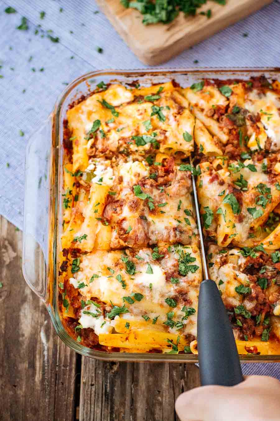 A woman is photographed from the front view as she is cutting into a freshly baked pasta bake