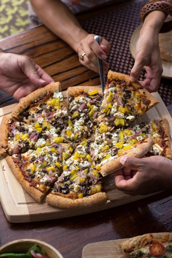 A person cutting slices of Whole-Wheat Pizza Crust and another person holding the pizza