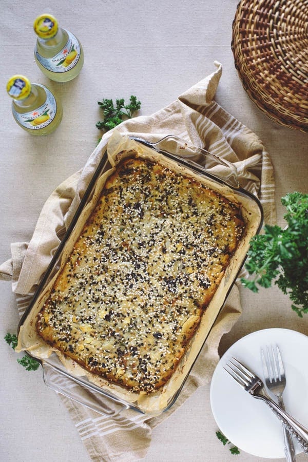 A Savory Feta Cheese and Kalamata Olive Cake in a baking dish on a napkin with 2 sparkling beverages and a basket on the side