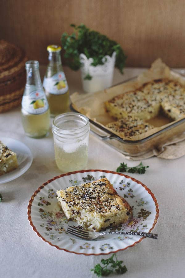 Single of Savory Feta Cheese and Kalamata Olive Cake with a plate of olive cake in the background and 2 beverages