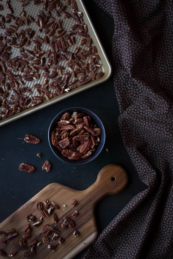 Chopped pecans on a cutting board, chopped pecans in a bowl and chopped pecans on a baking sheet with a decorative napkin on the side