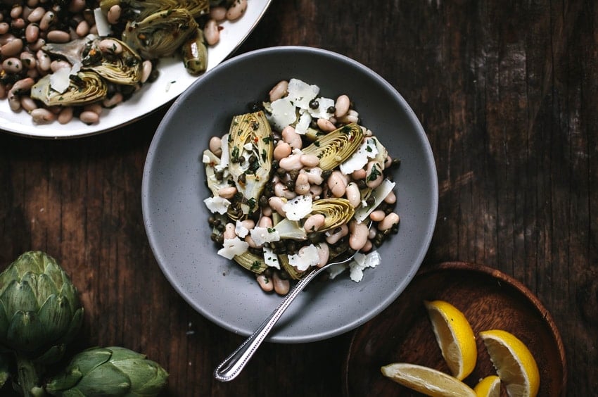 Bowl of Baby Artichoke Salad with White Beans and Manchego with lemons and artichokes on the side