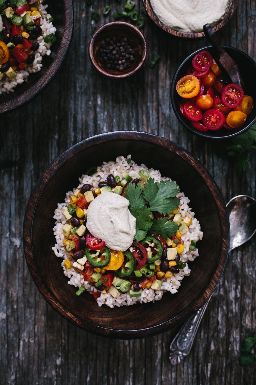 A large bowl of vegan burrito bowl topped off with homemade chipotle cashew sauce is photographed from the top view alongside with some of the ingredients.