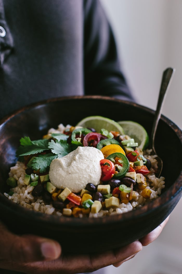 A man is holding a large bowl of Vegan Burrito Bowl topped off with Cashew Chipotle Cream Sauce photographed from the front view.
