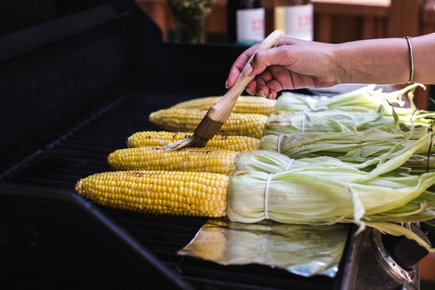 A woman is brushing corn on the grill with butter 