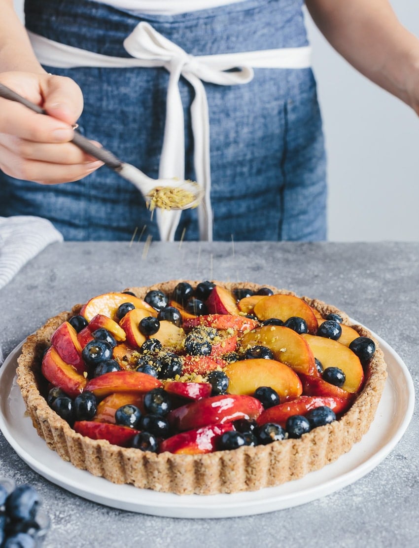 Person using a spoon to sprinkle crumbled pistachio over Peach and Blueberry Tart with Walnut Crust