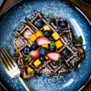 Brioche waffle topped off with fresh fruit on a plate with a fork on the side.