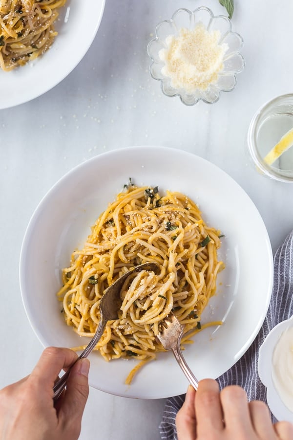 Overhead view of a person preparing to Pumpkin Creme Fraiche Pasta with Sage with a small bowl of cheese and a glass of lemon water