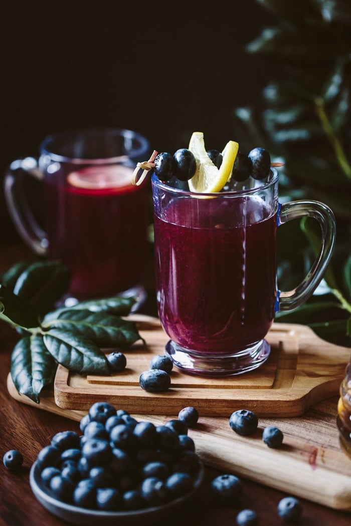 Close up view of a glass of Honey Sweetened Blueberry Hot Toddy garnished with a slice of lemon and blueberries and another hot toddy