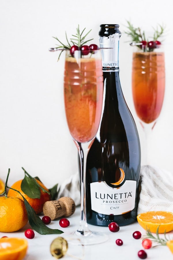 A bottle of Prosecco  with 2 flutes of Cocktails with 2 clementines, cork, and cranberries