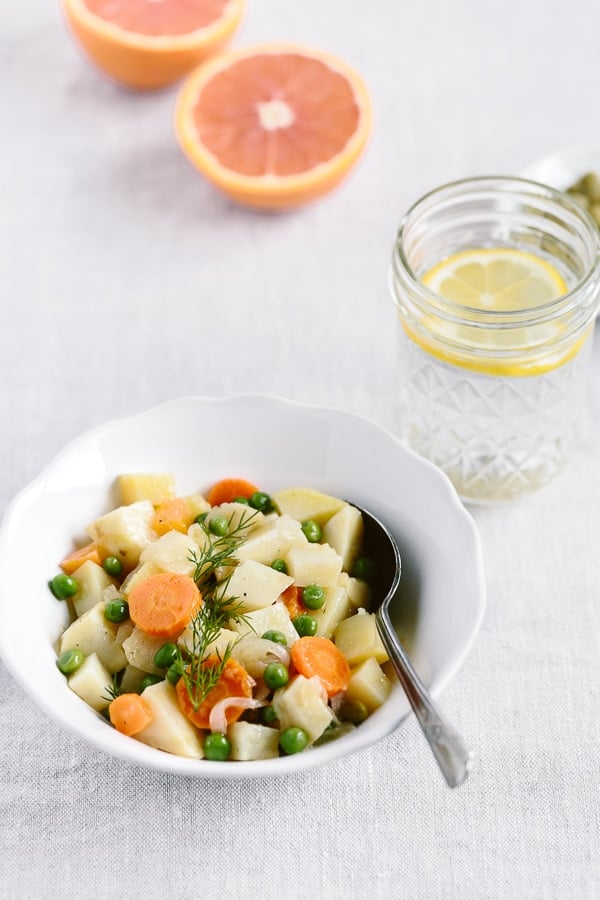 A bowl of One-Pot Citrusy Winter Root Vegetables with a spoon and a glass of lemon water and cut oranges