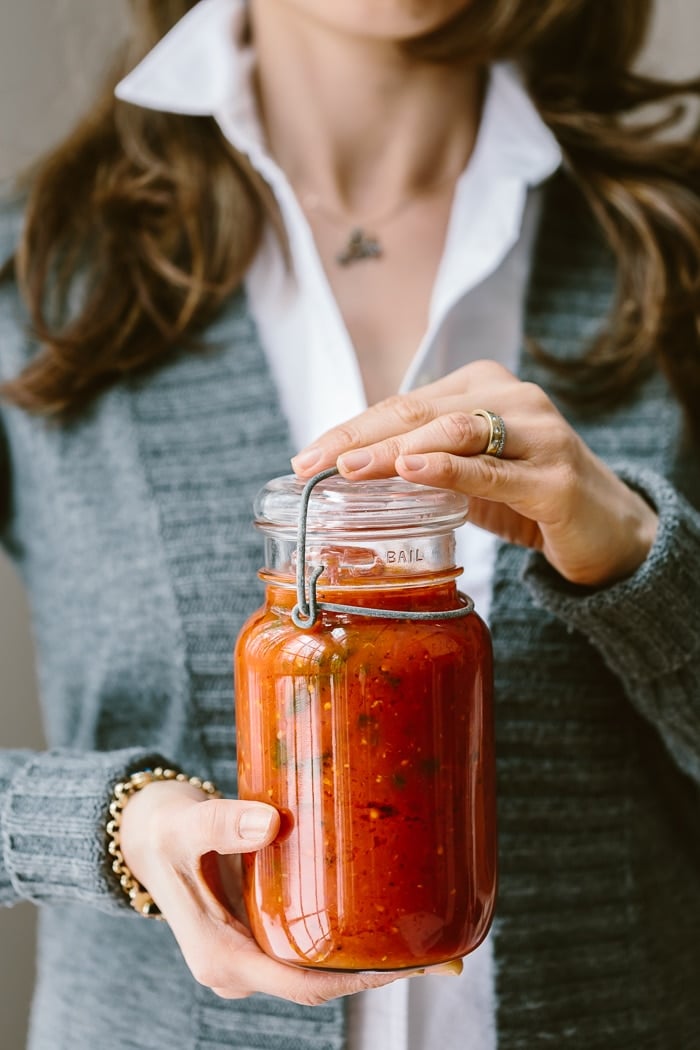 A woman is holding a jar of homemade tomato basil sauce photographed from the front view.