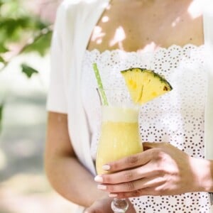 A woman is holding a glass of Pina Colada Smoothie: Frozen pineapple chunks blended with coconut milk, dates and nutmeg for a healthy morning smoothie.