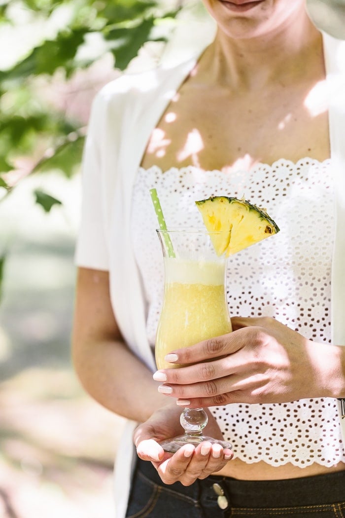 A woman is holding a glass of Pina Colada Smoothie: Frozen pineapple chunks blended with coconut milk, dates and nutmeg for a healthy morning smoothie.