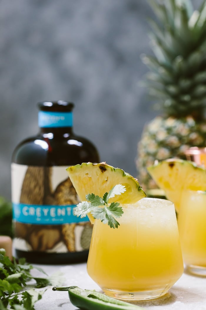 A glass of Spicy Pineapple Mezcalita with Jalapenos garnished with cilantri and a wedge of pineapple