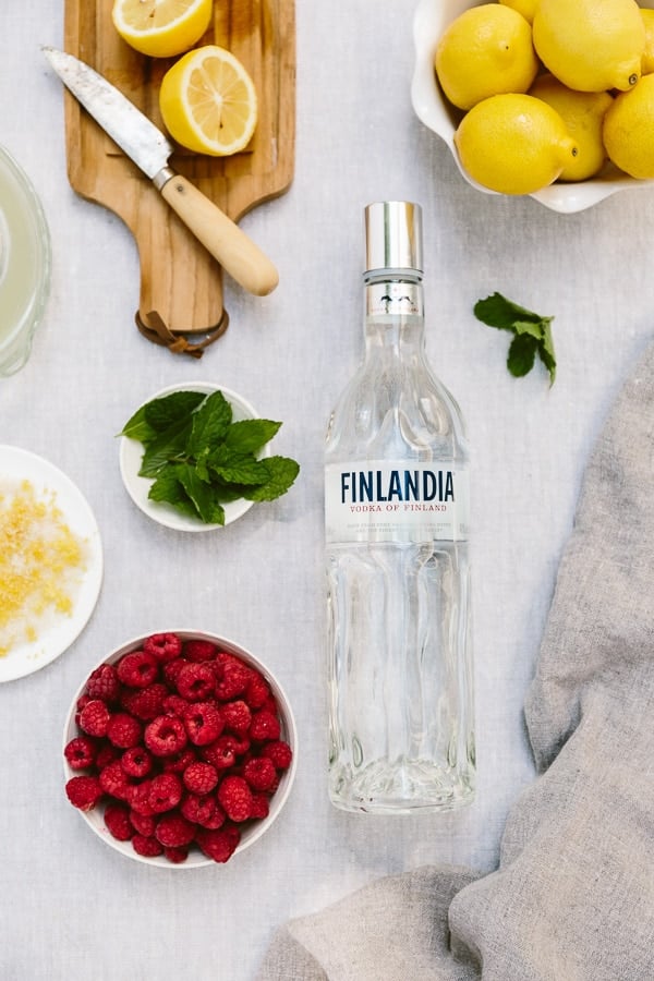 Ingredients for this recipe, lemon, mint, fresh raspberries, and vodka are laid out and photographed from the top.