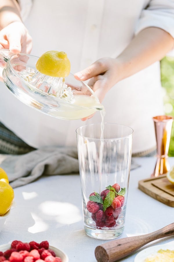 A woman is pouring lemon juice into a glass filled with raspberries and mint