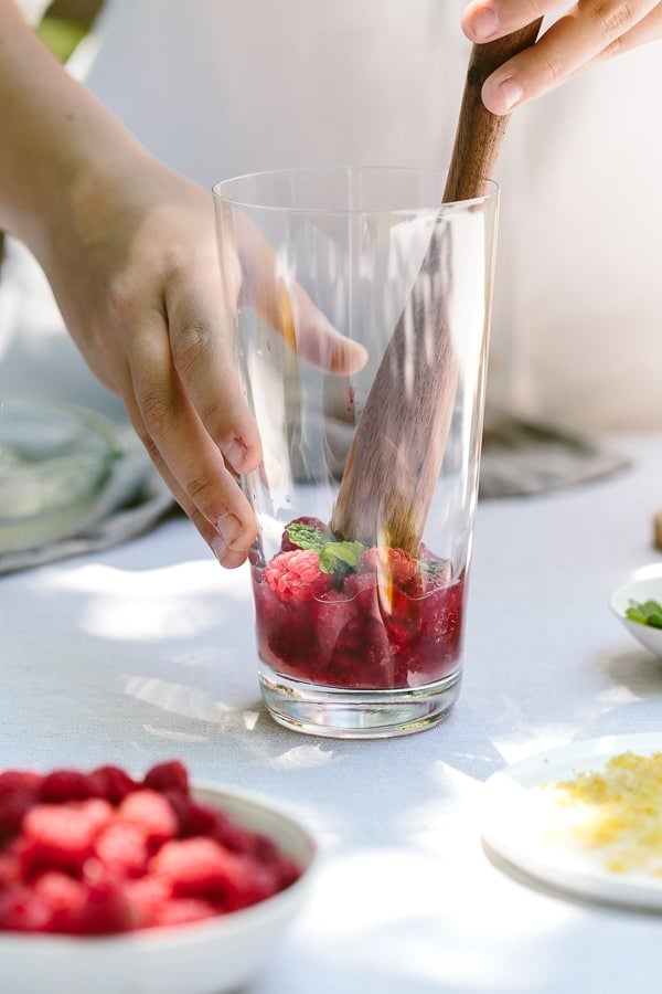 A woman is muddling raspberries and mint in a large glass as she preparing the cocktail