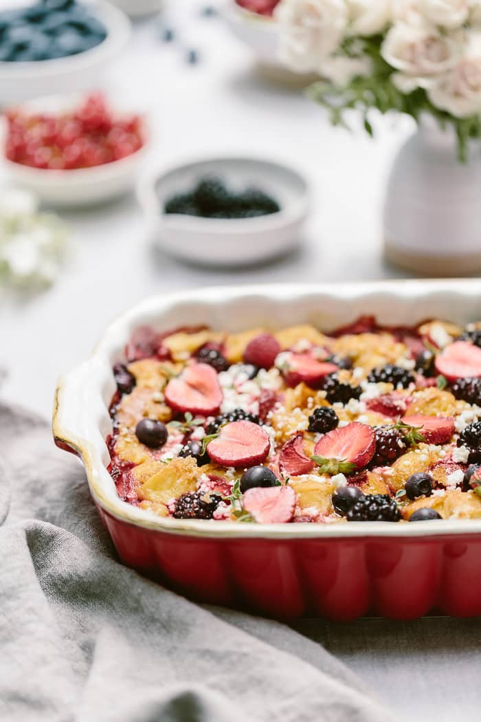 Goat cheese bread pudding with berries in a casserole dish with flowers in the background.