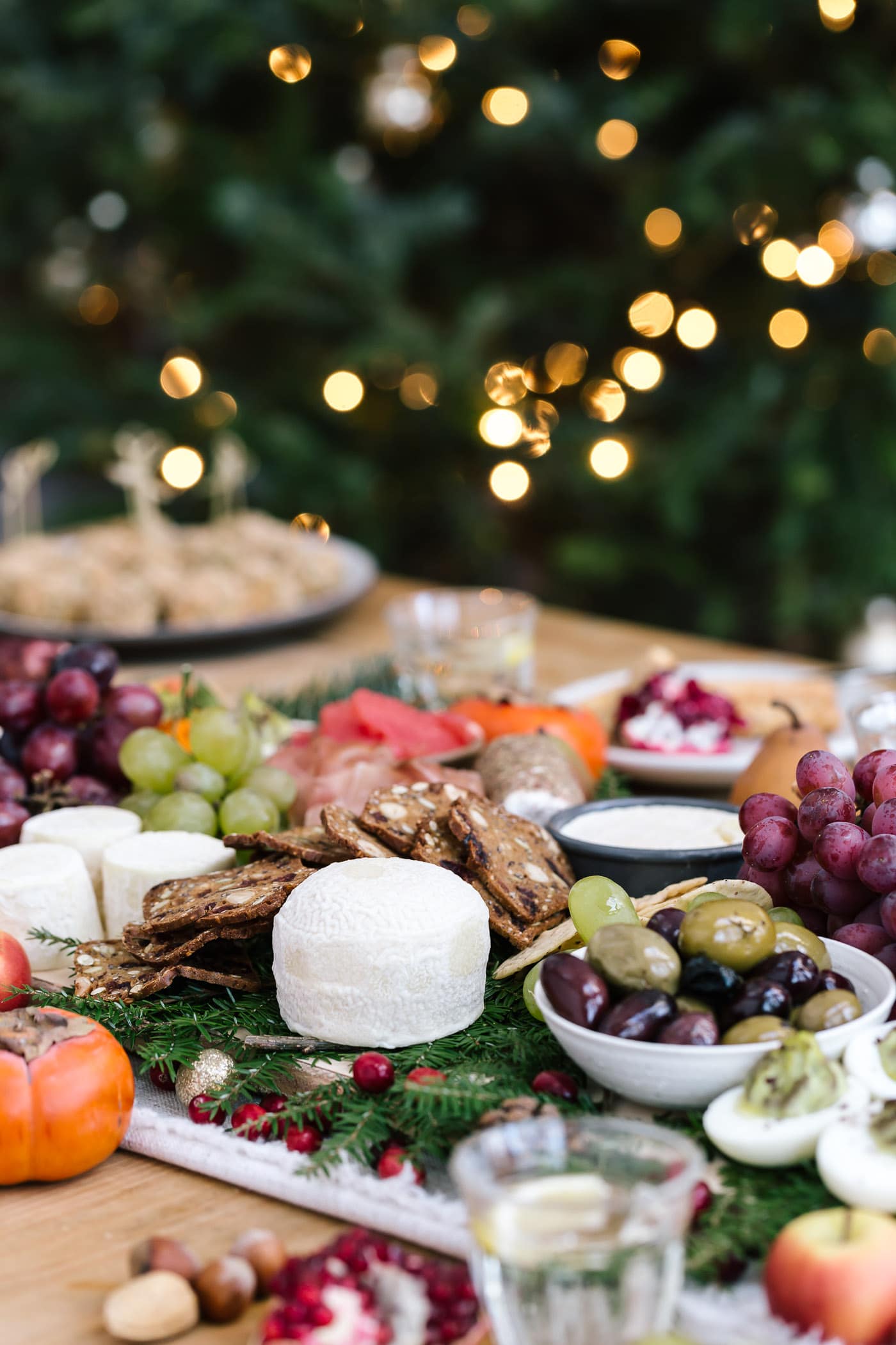 A table filled with cheese, baked goat cheese balls, fruit, olives and other edible items photographed with the Christmas tree in the background.