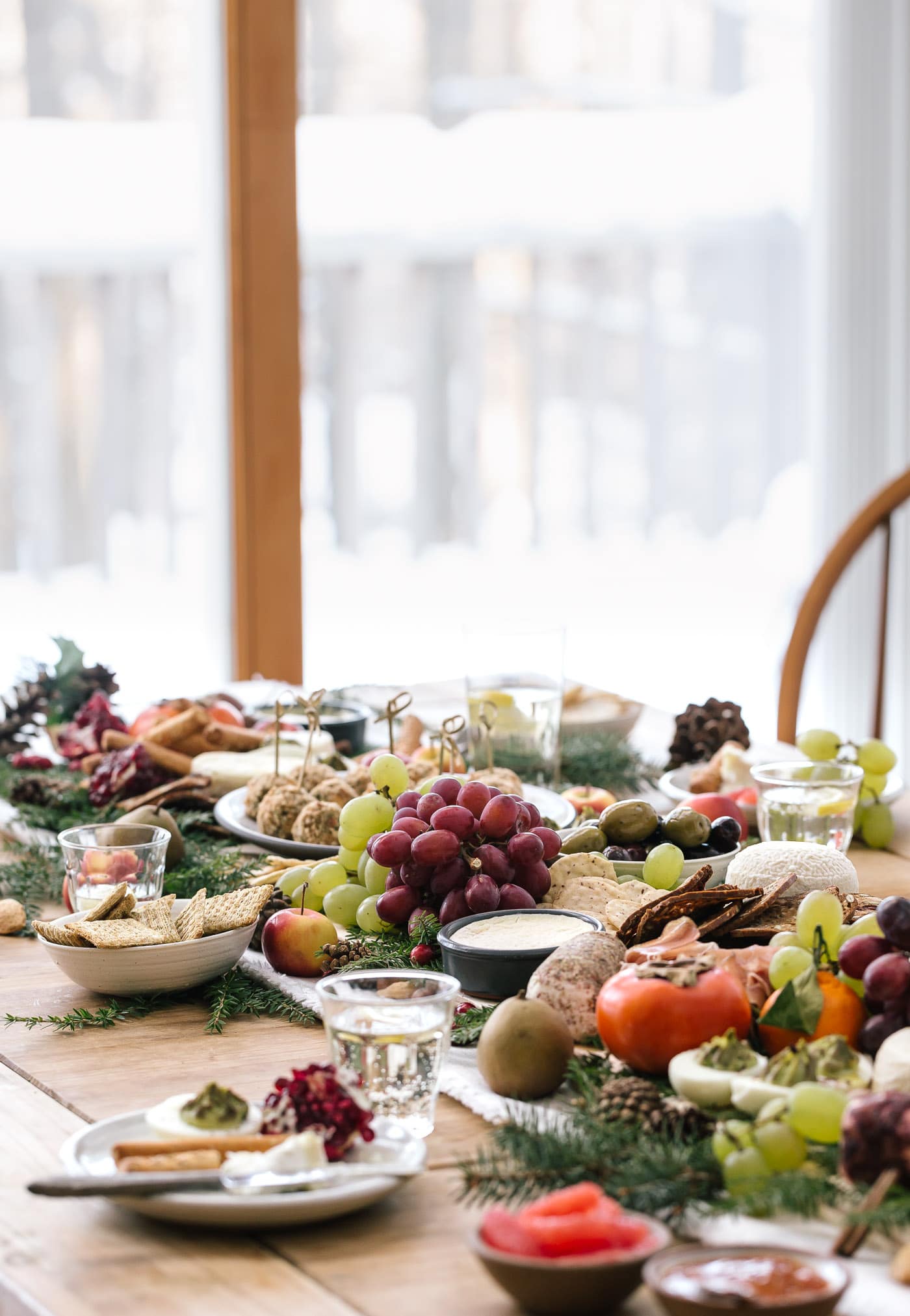 A table full of cheese, fruit and baked goat cheese balls are photographed from the front view with a snowy backdrop.