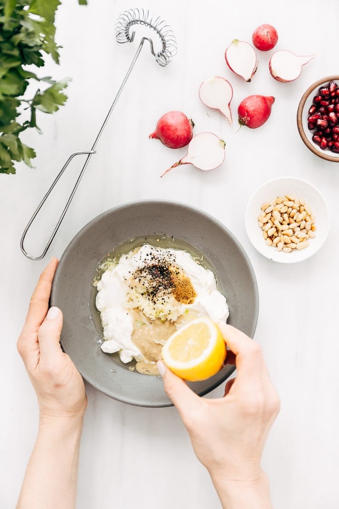 All ingredients for tahini yogurt sauce are placed in a bowl and a woman is squeezing lemon into the bowl is photographed from the top view.