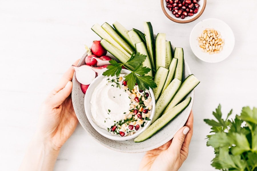 A bowl of tahini yogurt sauce surrounded with cucumbers and radishes is photographed as they are placed on the table by a woman from the top view.