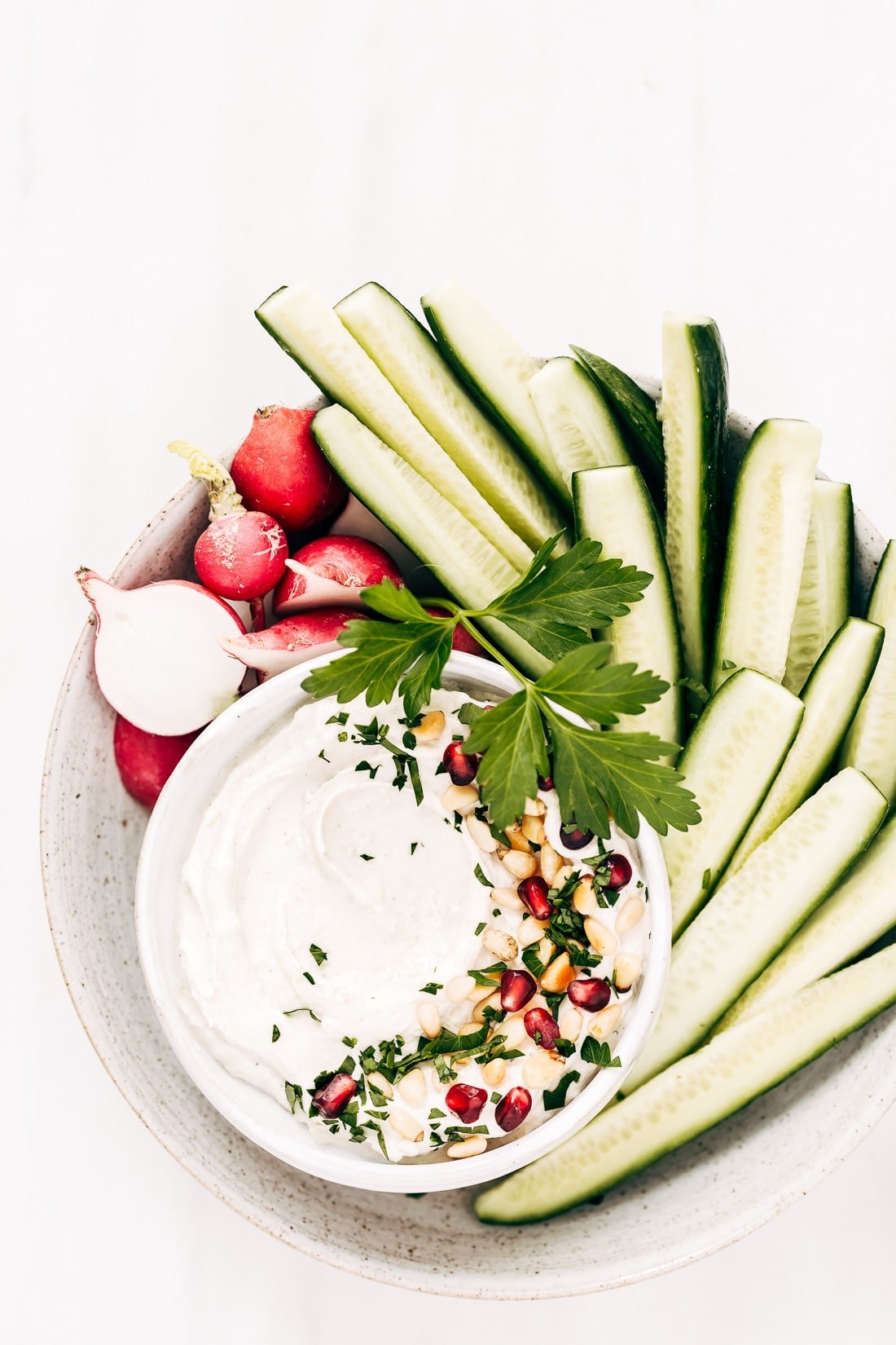 A bowl of tahini yogurt sauce surrounded with cucumbers and radishes is photographed from the top view.