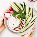 A bowl of tahini yogurt sauce surrounded with cucumbers and radishes is photographed from the top view.