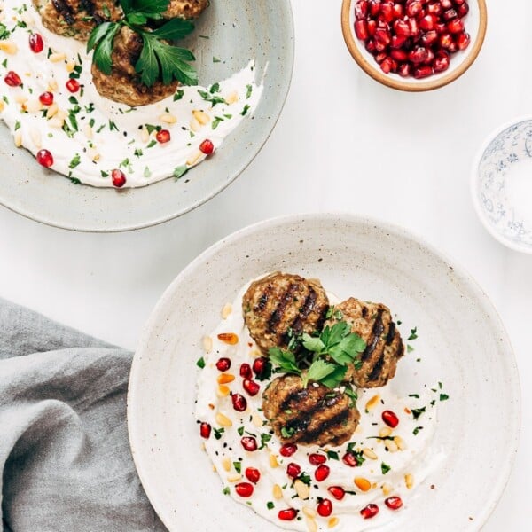 Two bowls filled with yogurt tahini sauce and Turkish meatballs garnished with pomegranate seeds and pine nuts.