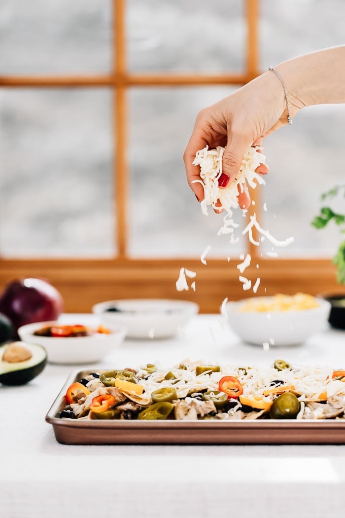 A woman is sprinkling cheese on a sheet pan of chicken nachos before it is baked.