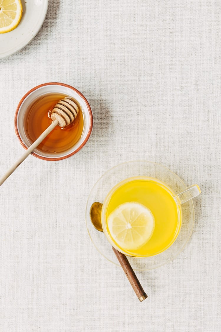 A glass cup of turmeric ginger tea with a lemon slice is photographed from the top view with a bowl of honey and sliced lemons.