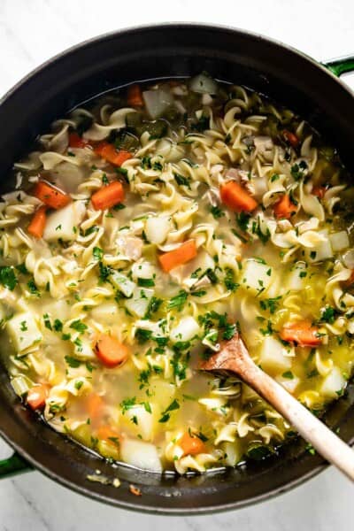 Chicken noodle soup with potatoes in a big pot with a wooden spoon on the side.