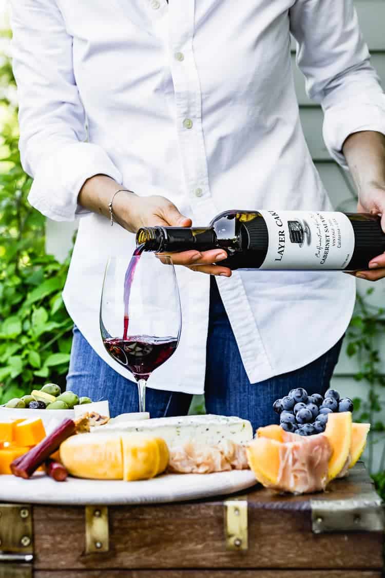 a woman is pouring wine in to a glass with a charcuterie and cheese board on the side