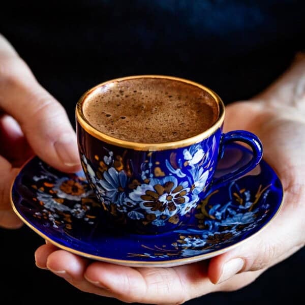 A woman holding a blue and gold Turkish coffee cup with thick foam on a matching saucer.