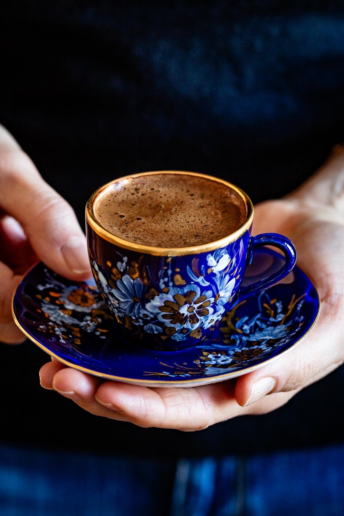 A woman holding a blue and gold Turkish coffee cup with thick foam on a matching saucer.