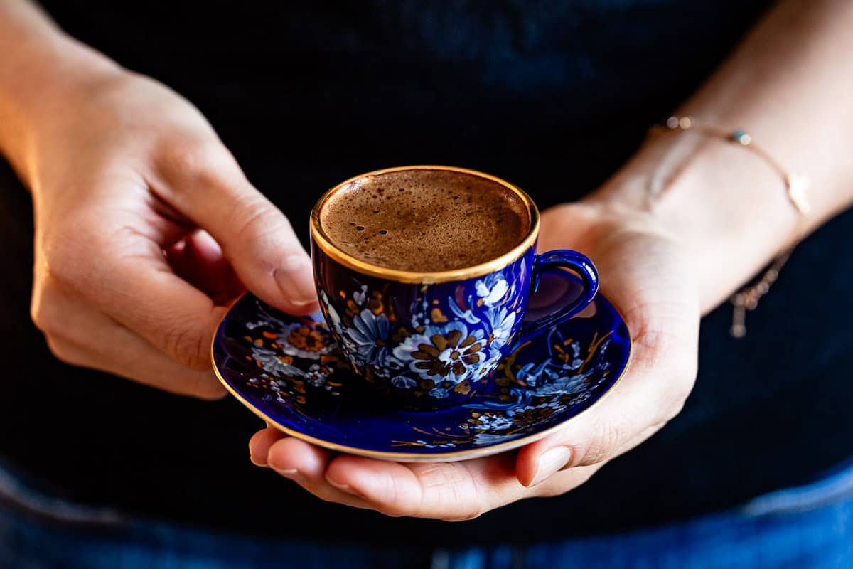 A woman holding a blue and gold Turkish coffee cup with thick foam on a matching saucer.