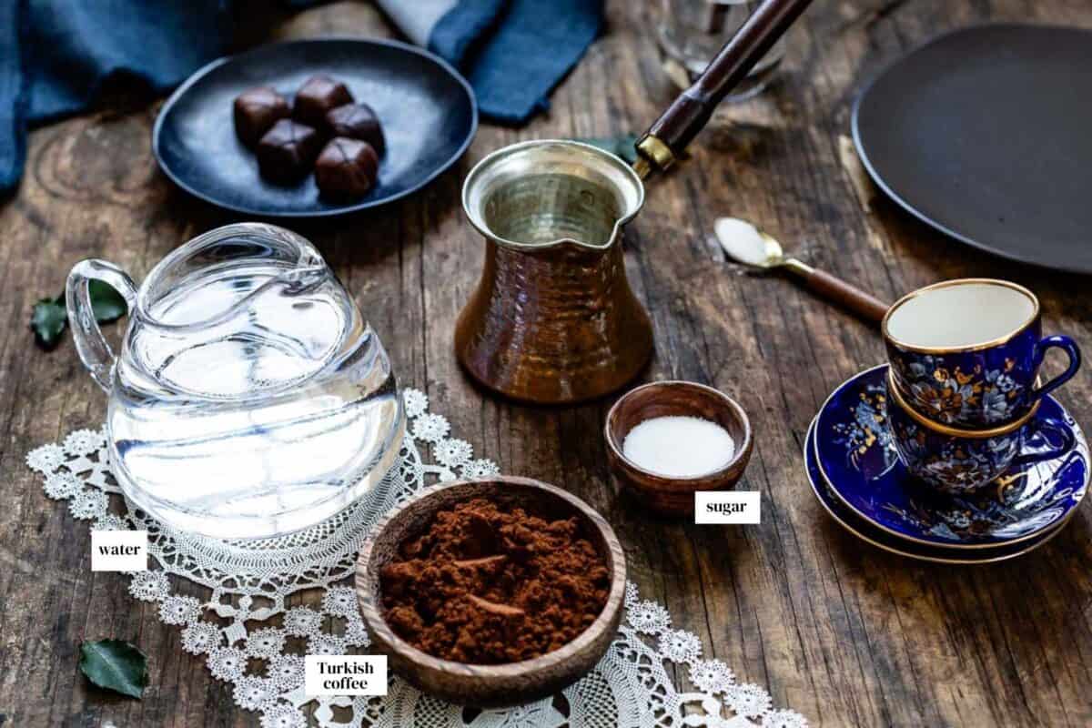 Turkish coffee ingredients laid out on a wooden surface including ground coffee, water, sugar, a copper cezve, and Turkish coffee cups.