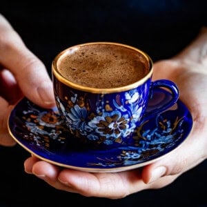 A woman holding a blue and gold Turkish coffee cup filled with thick foam on a matching saucer.
