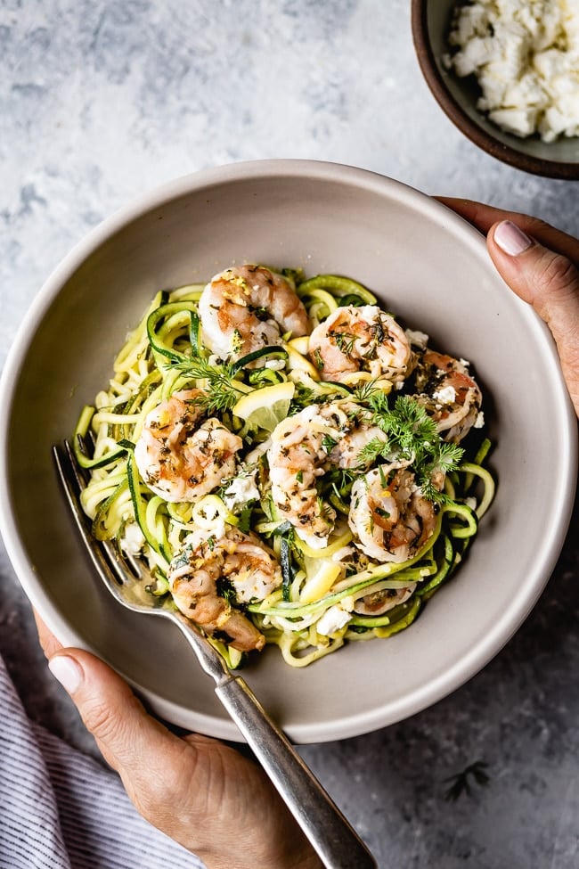 A bowl of Skinny Shrimp Scampi with Zucchini Noodles is photographed from the top view as a woman is holding it in her hands.