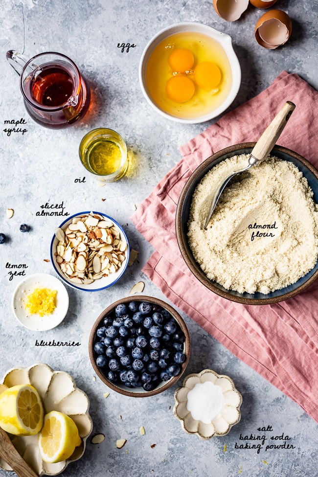 Ingredients (eggs, maple syrup, oil, almond flour, blueberries, lemon zest, etc.) are photographed from the top view.