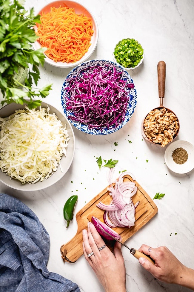 Ingredients: Carrots, red cabbage. green cabbage, jalapeno, celery seed, walnuts, and red onion as it is being sliced by a woman are photographed all together.