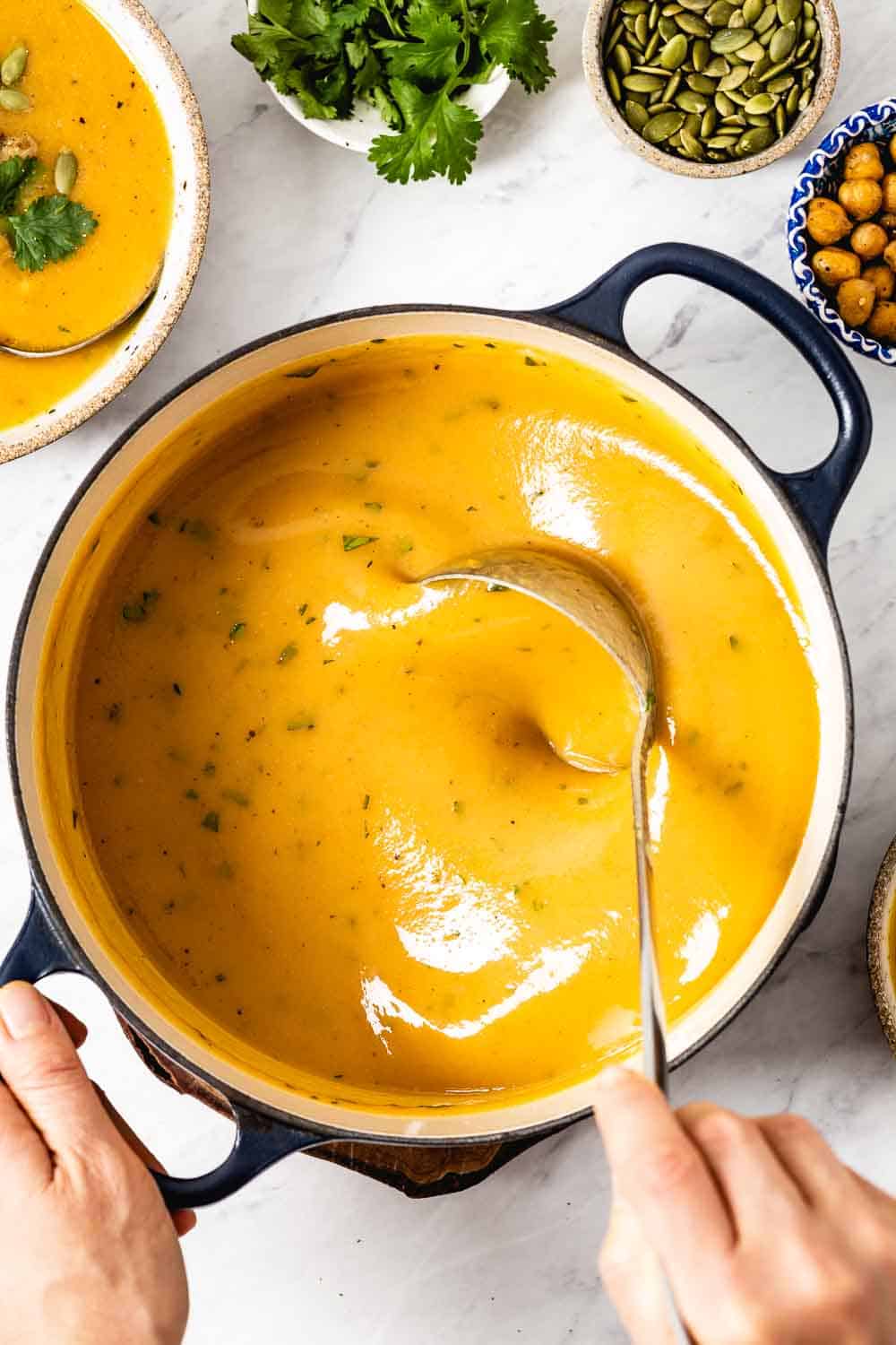 Creamy and Light Vegan Sweet Potato Carrot Ginger Soup is photographed from the top view in the pot as a woman is stirring it.