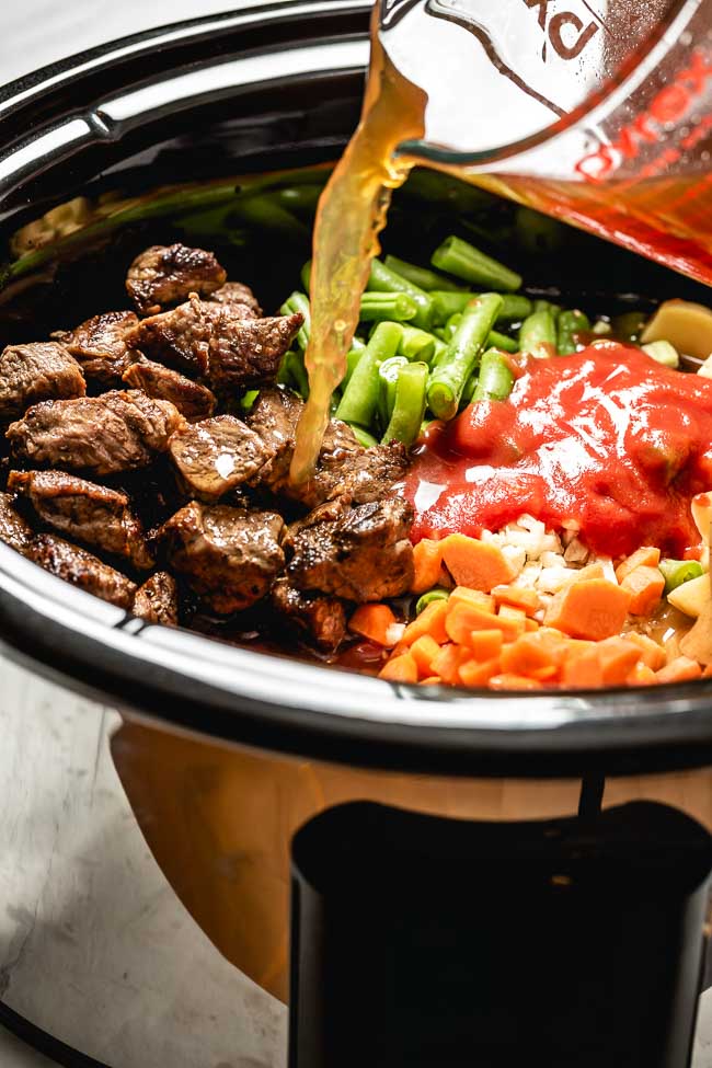 A crockpot filled with vegetables and beef is photographed as a woman is pouring beef broth into the mixture from the front view.