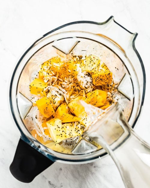 Mango Turmeric Ginger Smoothie ingredients are placed in a blender (Vitamix) and photographed from the top view as a woman is filling it with coconut water