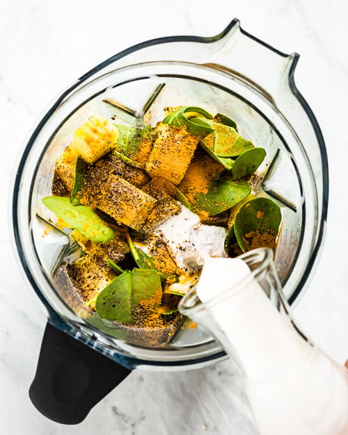 Turmeric Green Smoothie ingredients are placed in a blender and photographed from the top view as a woman is pouring almond milk into the blender