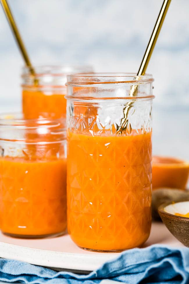 A jar filled with Orange ginger turmeric juice is photographed from a close up with a few other glasses in the background from the front view.