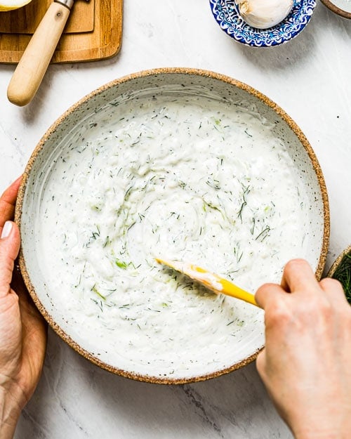 Yogurt with cucumber, garlic, and dill is being mixed by a woman
